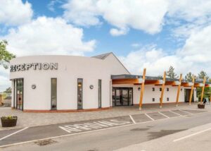 Contemporary new build reception building at Grange Leisure Holiday Park, designed by a Norwich-based architect. This welcoming structure features a curved rendered facade with full-height glazing, paired with a dynamic timber canopy supported by angled wooden columns. The modern design blends white render, timber, and glass for a bright and inviting arrival experience, emphasizing natural materials and a connection to the surrounding landscape.